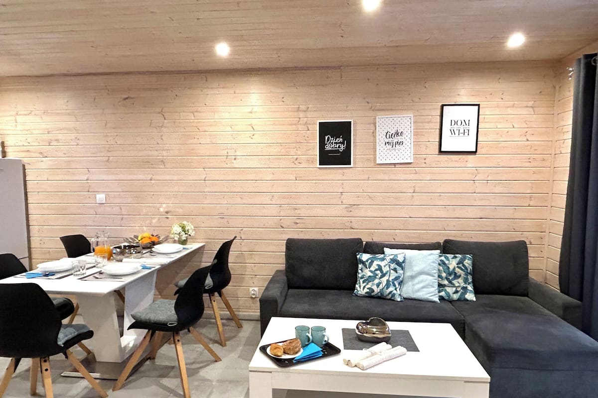 Una sala moderna con un sofá, una mesa de centro y un comedor. Hay cuadros enmarcados en la pared y un refrigerador al fondo.