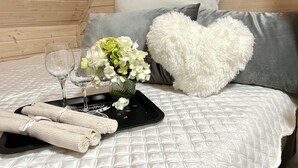Een netjes opgemaakt bed met een gebogen witte sprei, een zacht kussen in de vorm van een hart, een vaas met witte bloemen, twee lege wijnglazen en een dienblad met opgerolde handdoeken.