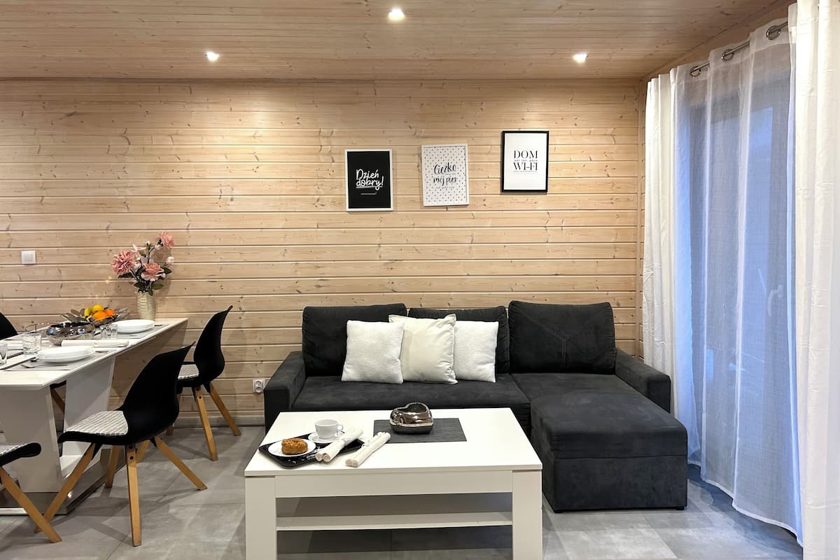 Una sala de estar moderna con techo de madera, un sofá negro con cojines blancos, una mesa de centro blanca y un comedor con mesa y sillas de madera.