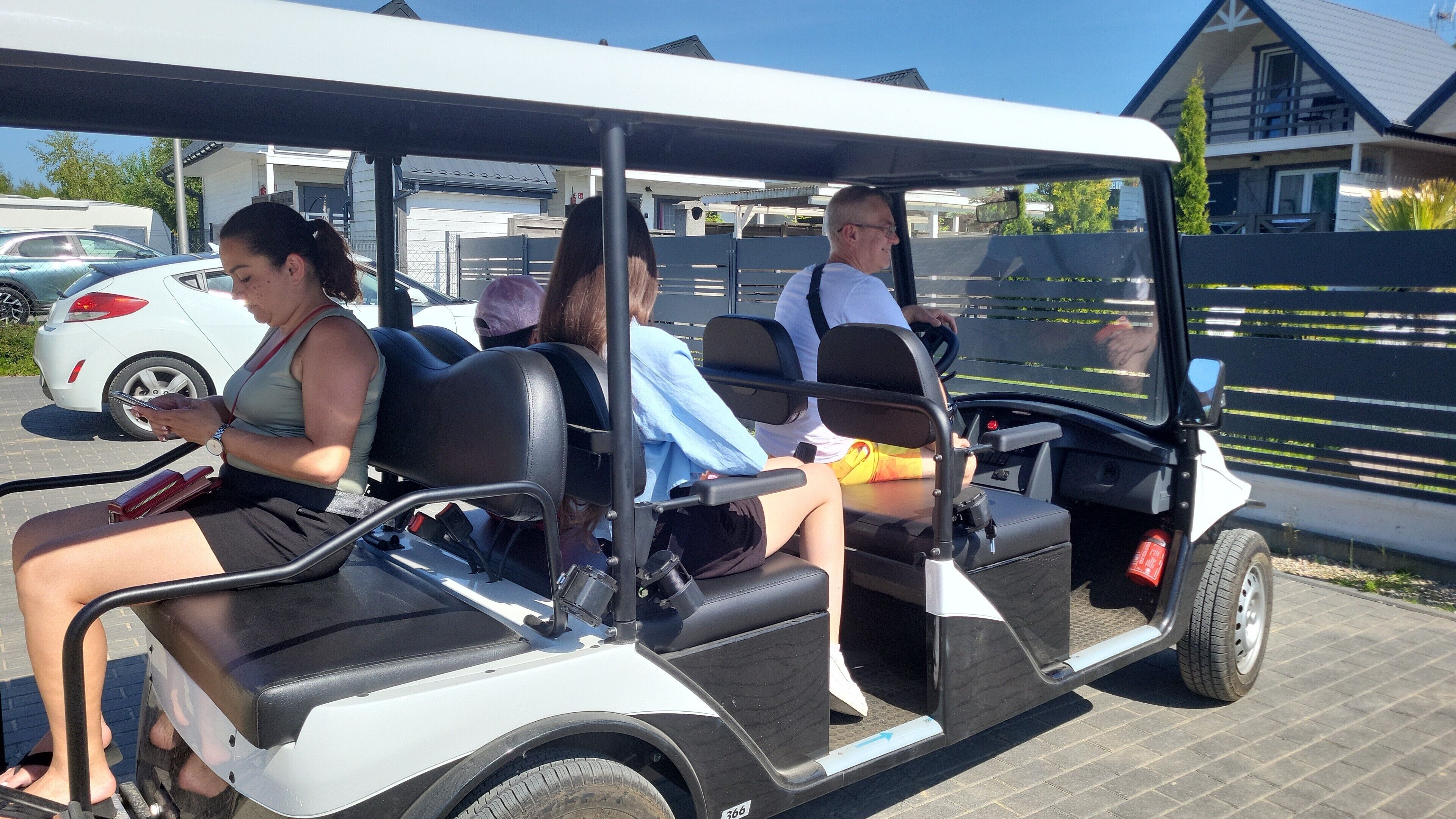 A golf cart with passengers, parked in a parking lot with buildings in the background.