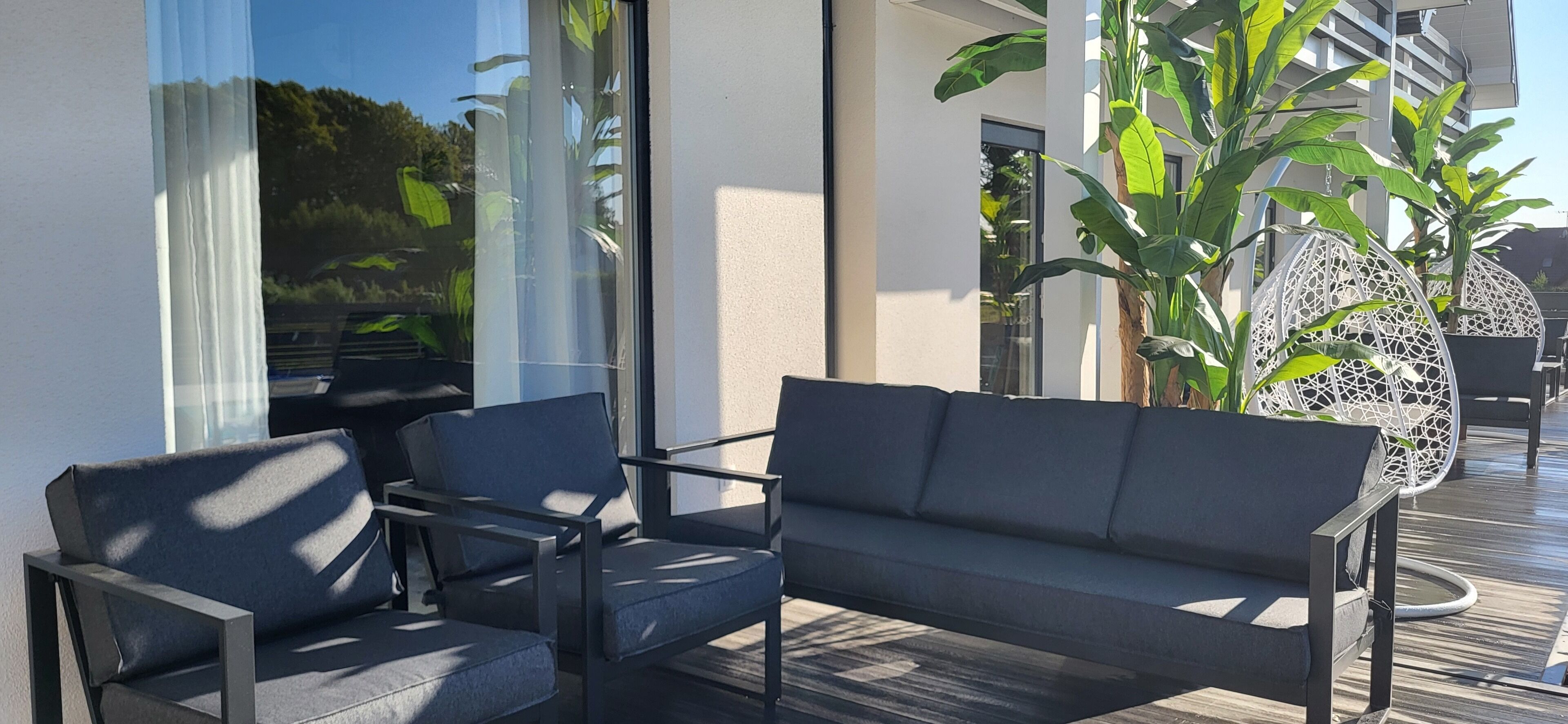 Un espace extérieur moderne aménagé avec un canapé et des fauteuils, de grandes fenêtres et des plantes en pot.