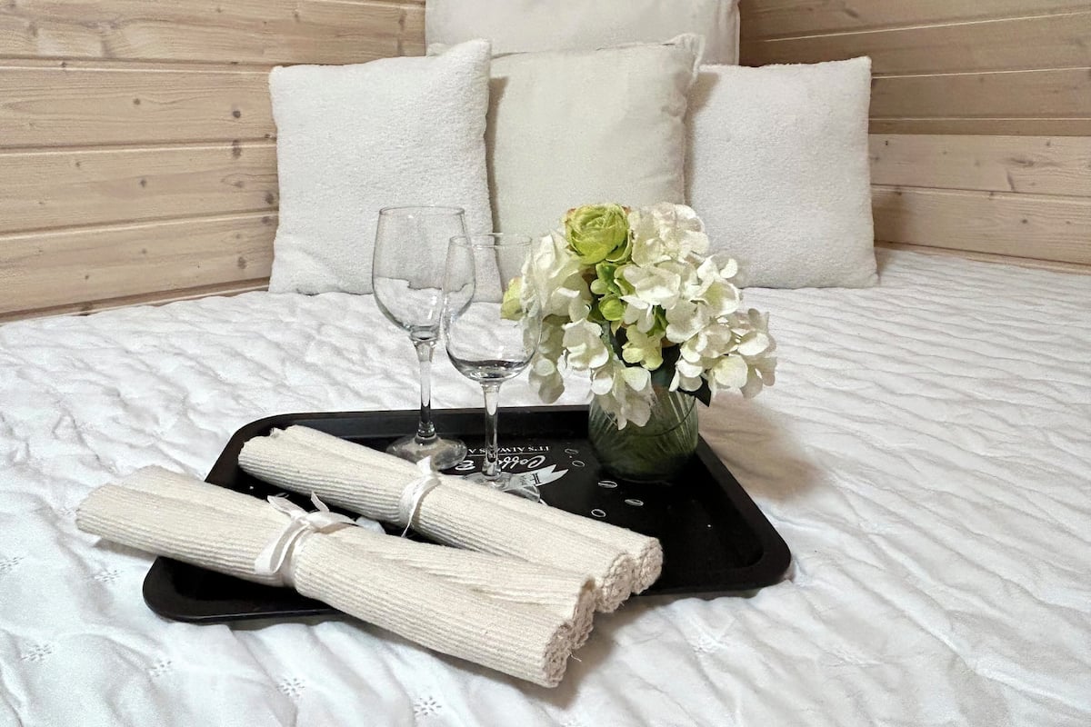 Una cama bien hecha con sábanas blancas, una bandeja con un jarrón de flores blancas y dos copas de vino vacías.
