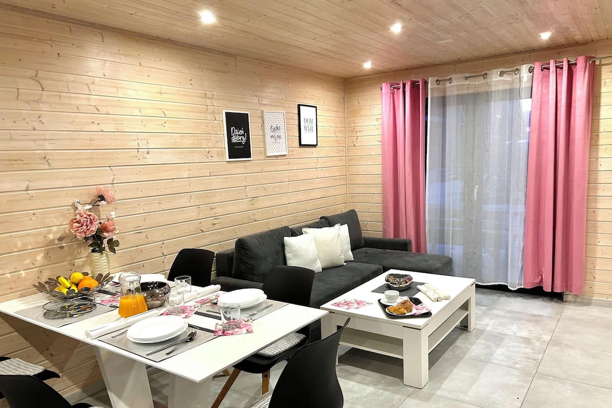 Una sala de estar moderna con una pared de madera, un sofá gris, una mesa de comedor con platos, vasos y un jarrón de flores.