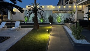 Een goed verlicht terras buiten, met palmbomen, een pad en zitplaatsen.