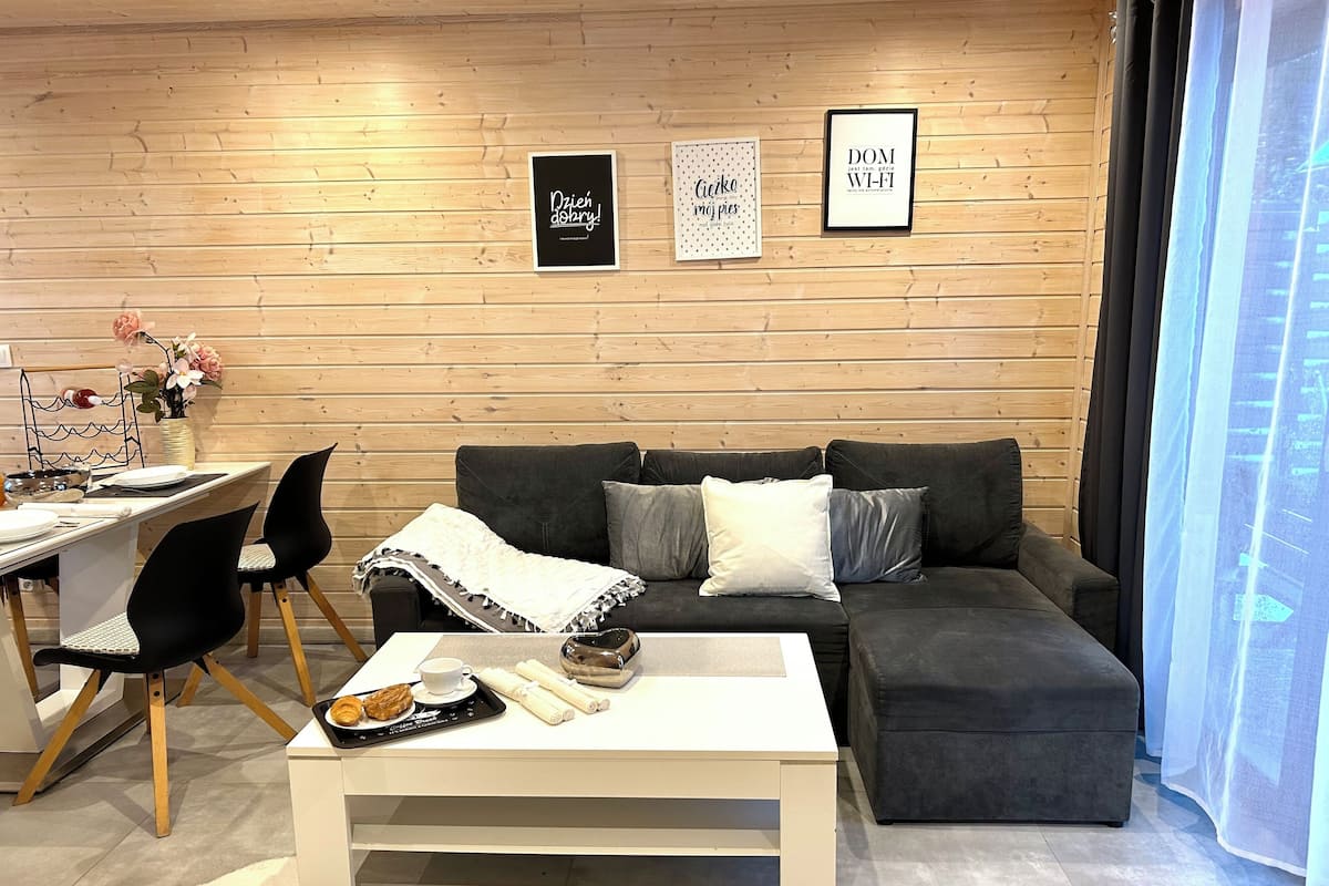 Una sala de estar moderna con una pared de madera, un sofá gris, una mesa de centro blanca y un comedor con una mesa blanca y sillas negras.