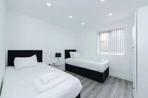 A hotel room with two beds, a window with blinds, a bedside table with a lamp, and a wardrobe.