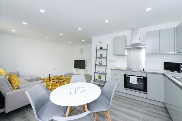 A modern kitchen with white cabinets and a stainless steel range hood, a dining area with a white table and grey chairs, and a grey sofa with yellow cushions.