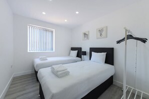 A hotel room with two beds, a window with blinds, a coat rack, and artwork on the wall. - Nottingham 2-BR Apartment w/ Parking (Gedling)