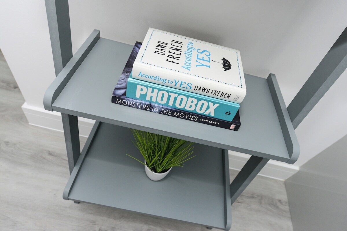 A grey metal shelf with a potted plant, holding books titled 'According to YES' and 'PHOTOBOX.'