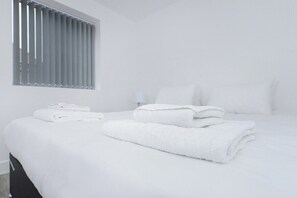 A hotel room with a bed, white linens, a window with blinds, and a nightstand with a lamp. - Apartment w/ Parking Close to Nottingham (Gedling)