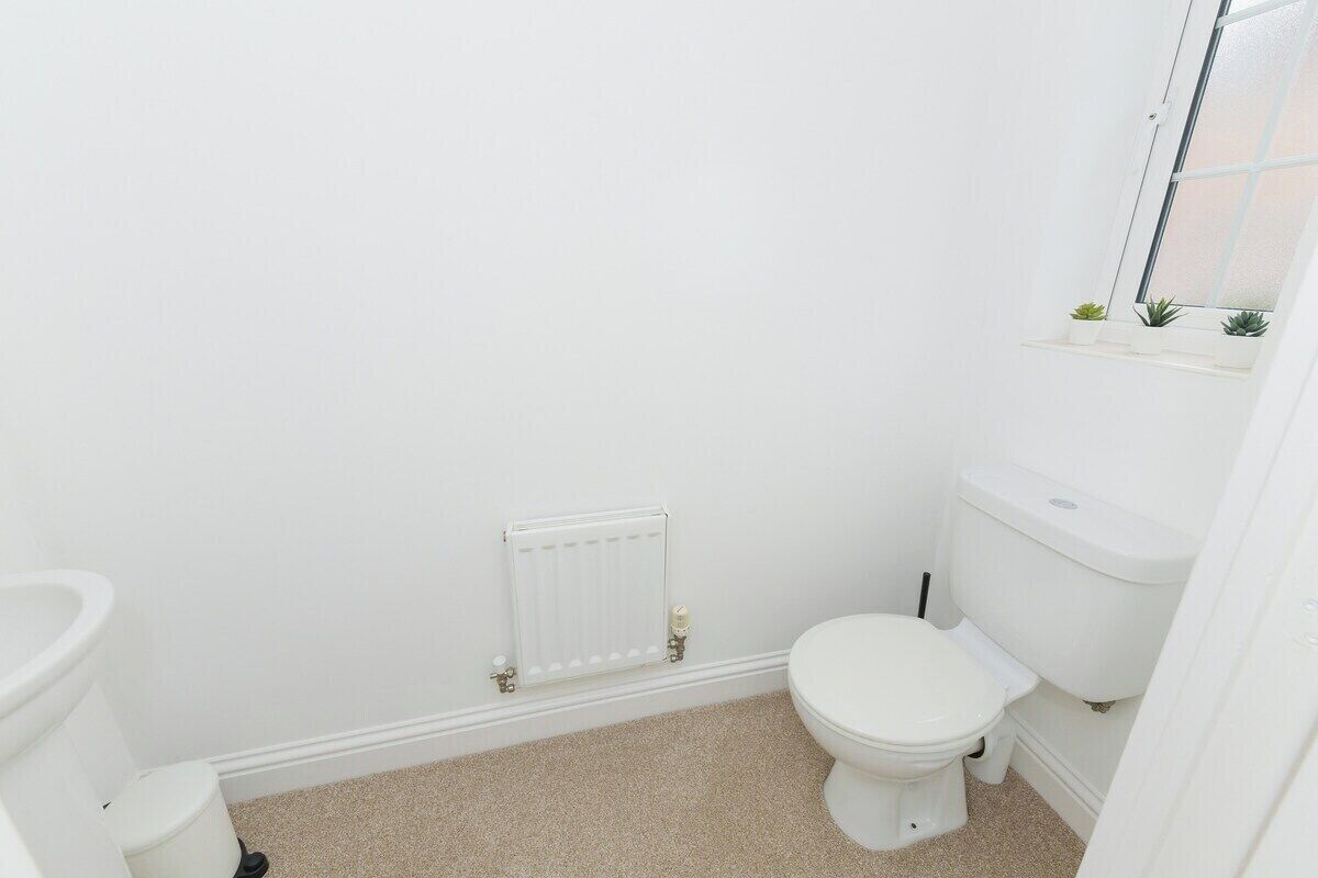 Une petite salle de bain blanche, avec des toilettes, un radiateur et une fenêtre agrémentée de quelques petites plantes.