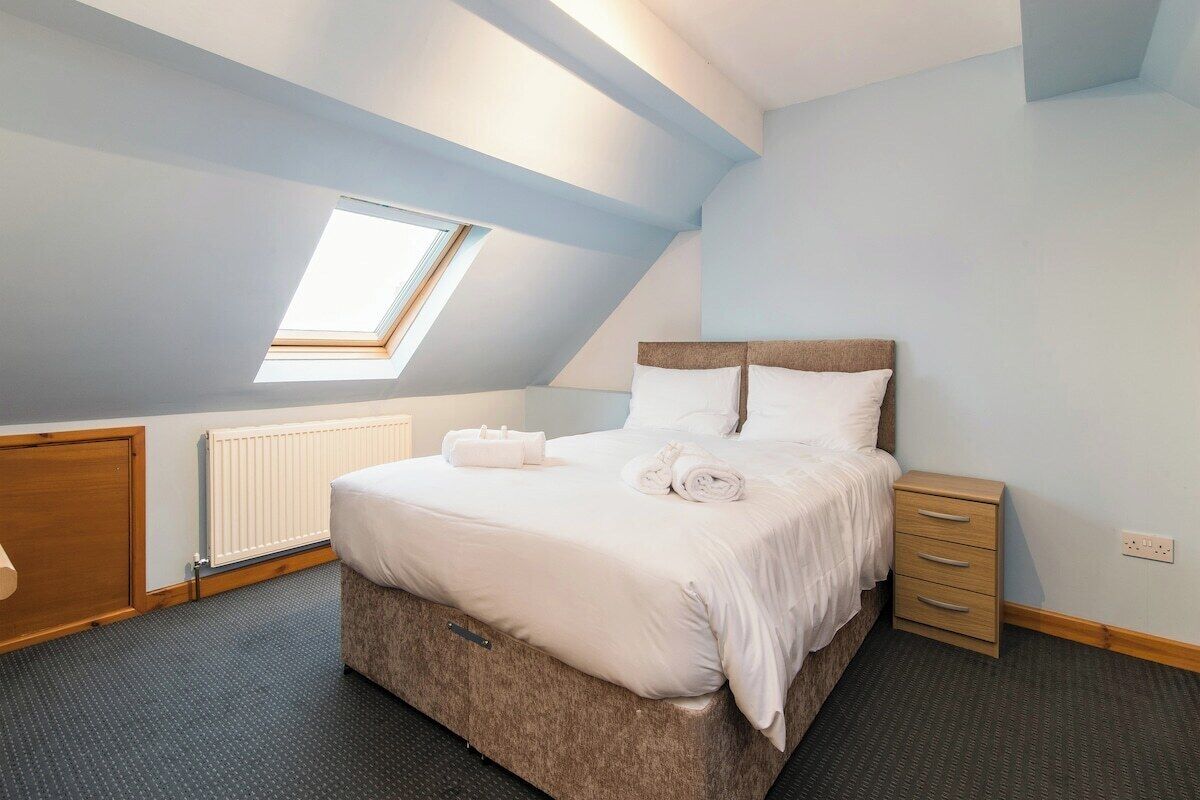A bedroom with a sloped ceiling, a skylight, a bed with white linens, a wooden nightstand, and a radiator.