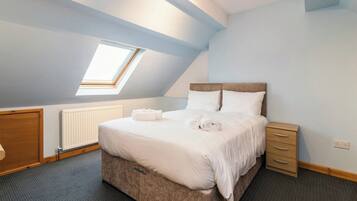 A bedroom with a sloped ceiling, a skylight, a bed with white linens, a wooden nightstand, and a radiator.