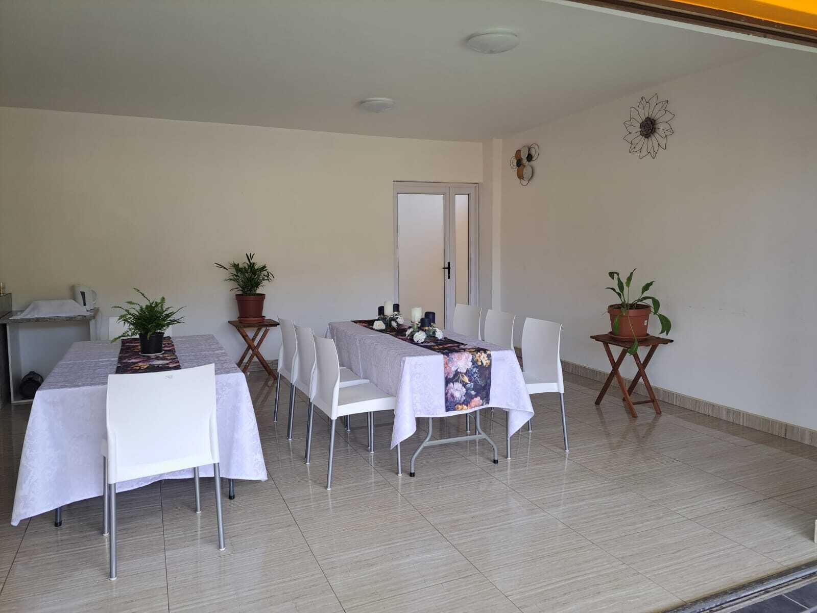 Un comedor con una mesa dispuesta para seis personas, rodeada de sillas blancas, plantas en macetas sobre mesas pequeñas y una puerta al fondo.