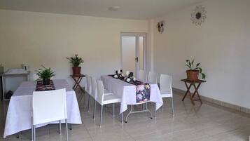 Un comedor con una mesa dispuesta para seis personas, rodeada de sillas blancas, plantas en macetas sobre mesas pequeñas y una puerta al fondo.