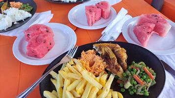 Een tafel met diverse gerechten, waaronder rijst, watermeloen, friet en een visgerecht.