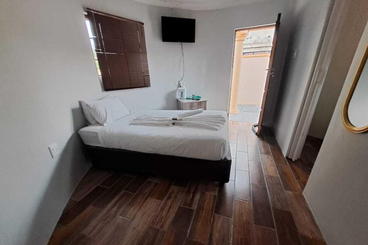 A bedroom with a bed, a TV mounted on the wall, a window with blinds, and a wooden floor.