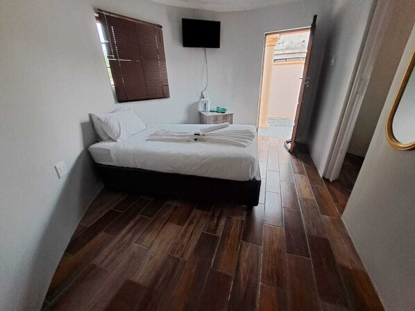 A bedroom with a bed, a TV mounted on the wall, a window with blinds, and a wooden floor.