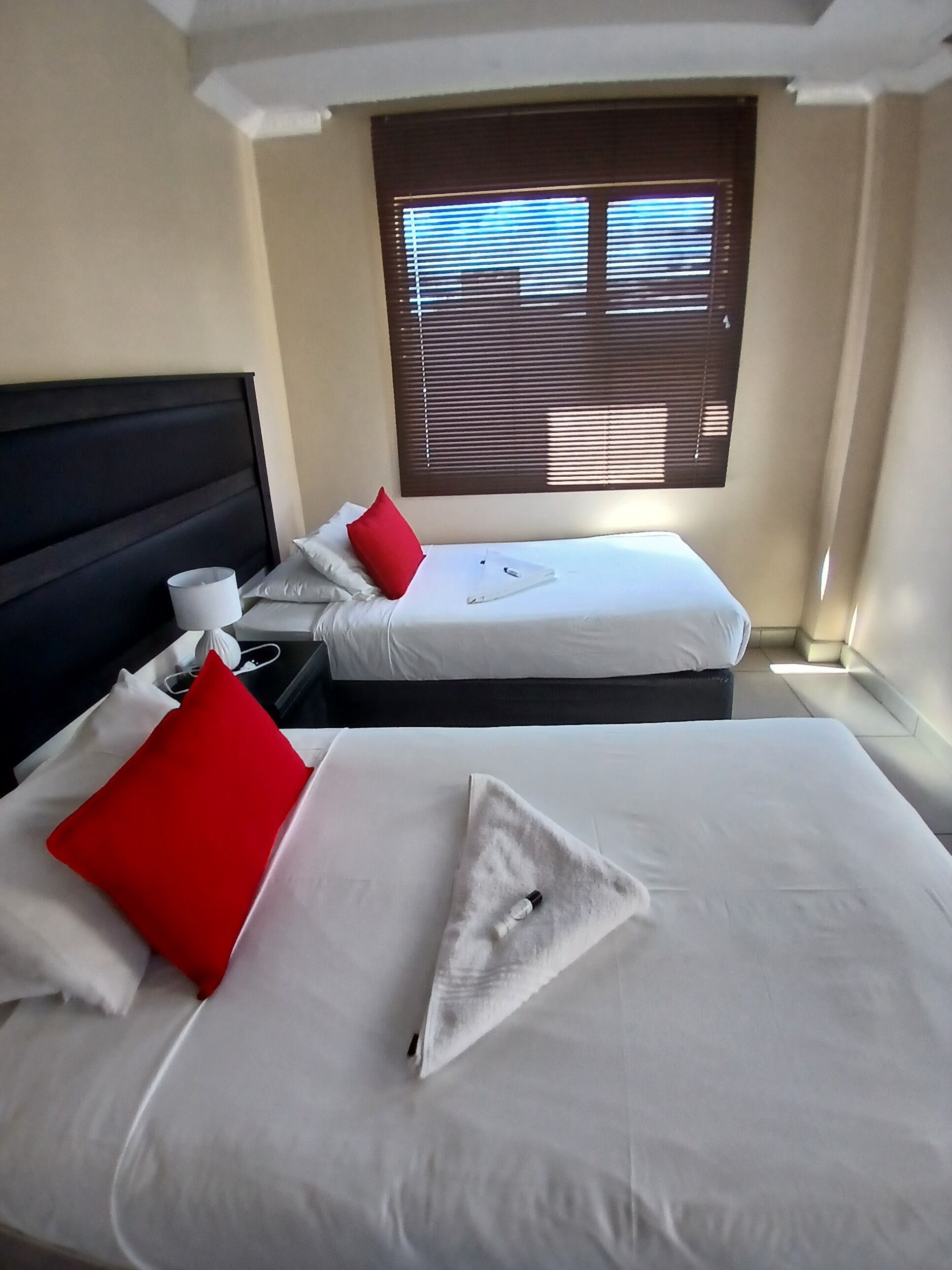 A hotel room with two beds, each with white bedding and red pillows, a nightstand with a lamp, and a window with blinds.
