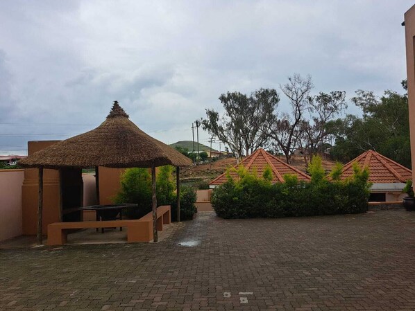 A courtyard with a thatched-roof gazebo, a brick pathway, and several buildings with red-tiled roofs. - Jate Guest House (Maseru)