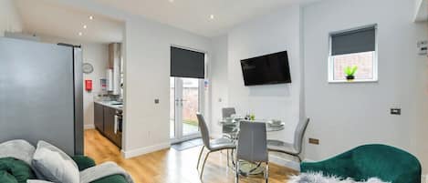 A modern living room with a sofa, a teal armchair, a dining area with a table and chairs, a kitchen in the background, and a TV mounted on the wall.