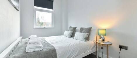 A bedroom with a single bed, a bedside table with a lamp, a window, and a wall-mounted power outlet.