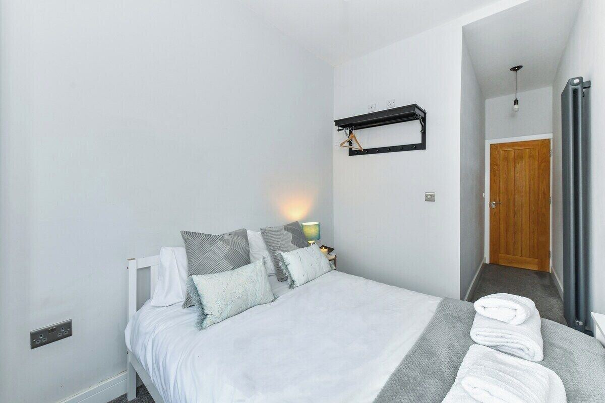 A bedroom with a bed, pillows, a bedside table with a lamp, a towel rack, and a door leading to another room.