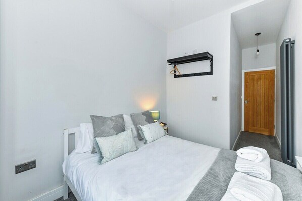 A bedroom with a bed, pillows, a bedside table with a lamp, a towel rack, and a door leading to another room.