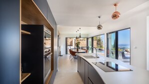 Een moderne keuken met een groot eiland, een eethoek met tafel en stoelen, en uitzicht op de omgeving door grote ramen.