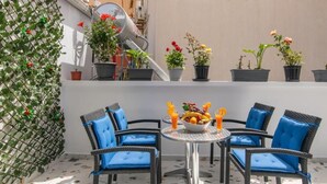 Um pátio com uma mesa redonda, quatro cadeiras azuis com almofadas, uma tigela com frutas e bebidas. Há plantas em vasos e uma trepadeira na parede.