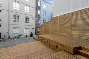 A modern outdoor deck with wooden steps leading up to a flat surface, surrounded by multi-story residential buildings with balconies and windows. - Swan Deluxe Studio for 4 guests (Reykjavík)