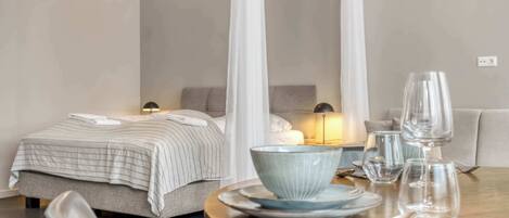 A hotel room with a bed, bedside table with a lamp, a dining table with plates, glasses, and cutlery.