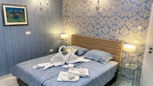 Un lit bien fait, recouvert d’une couvre-lit bleue à motifs, deux lampes fixées au mur, un tableau encadré et un papier peint à motifs.