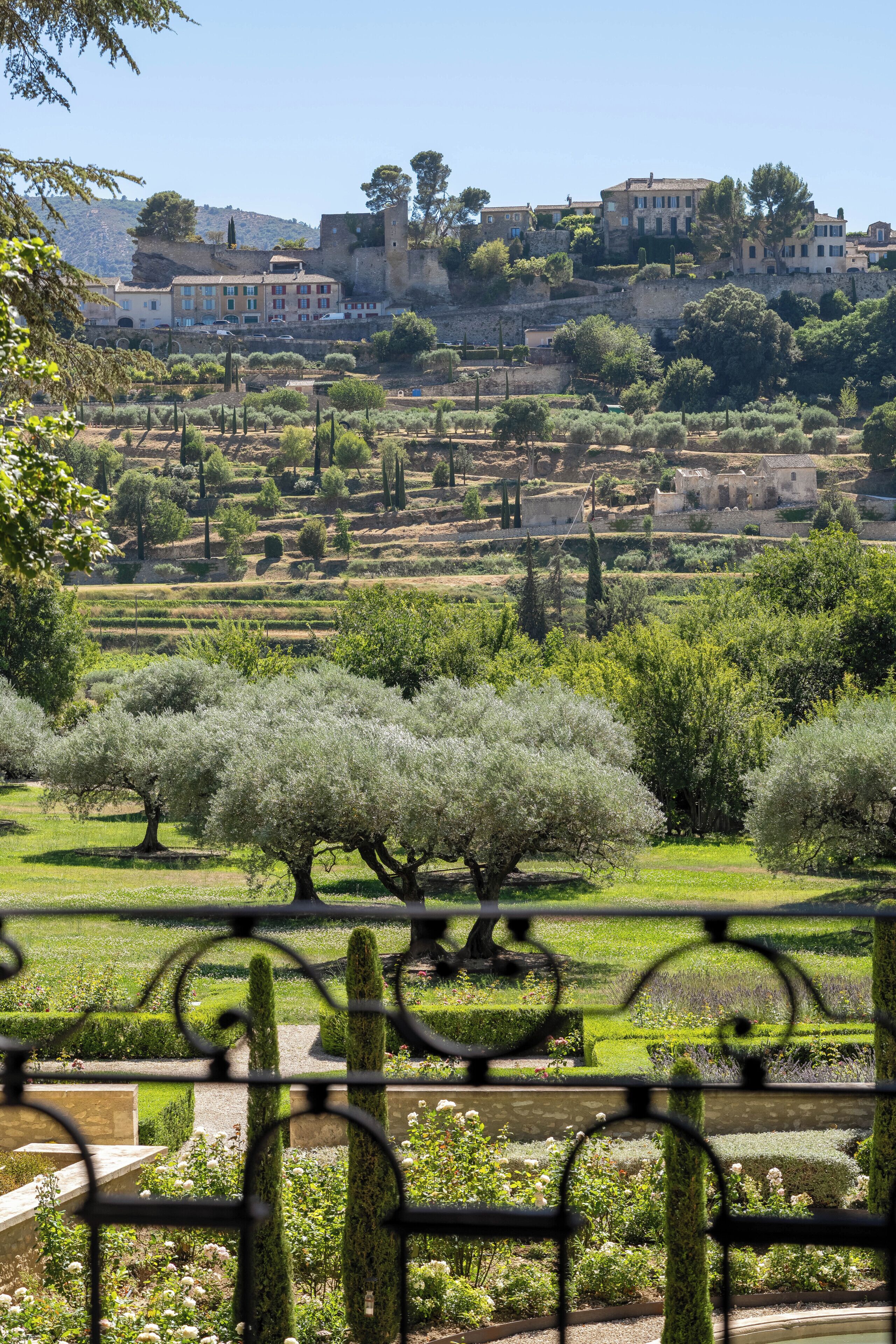A view of a historic town with buildings on a hillside, a well-maintained garden with cypress trees, and a clear sky.