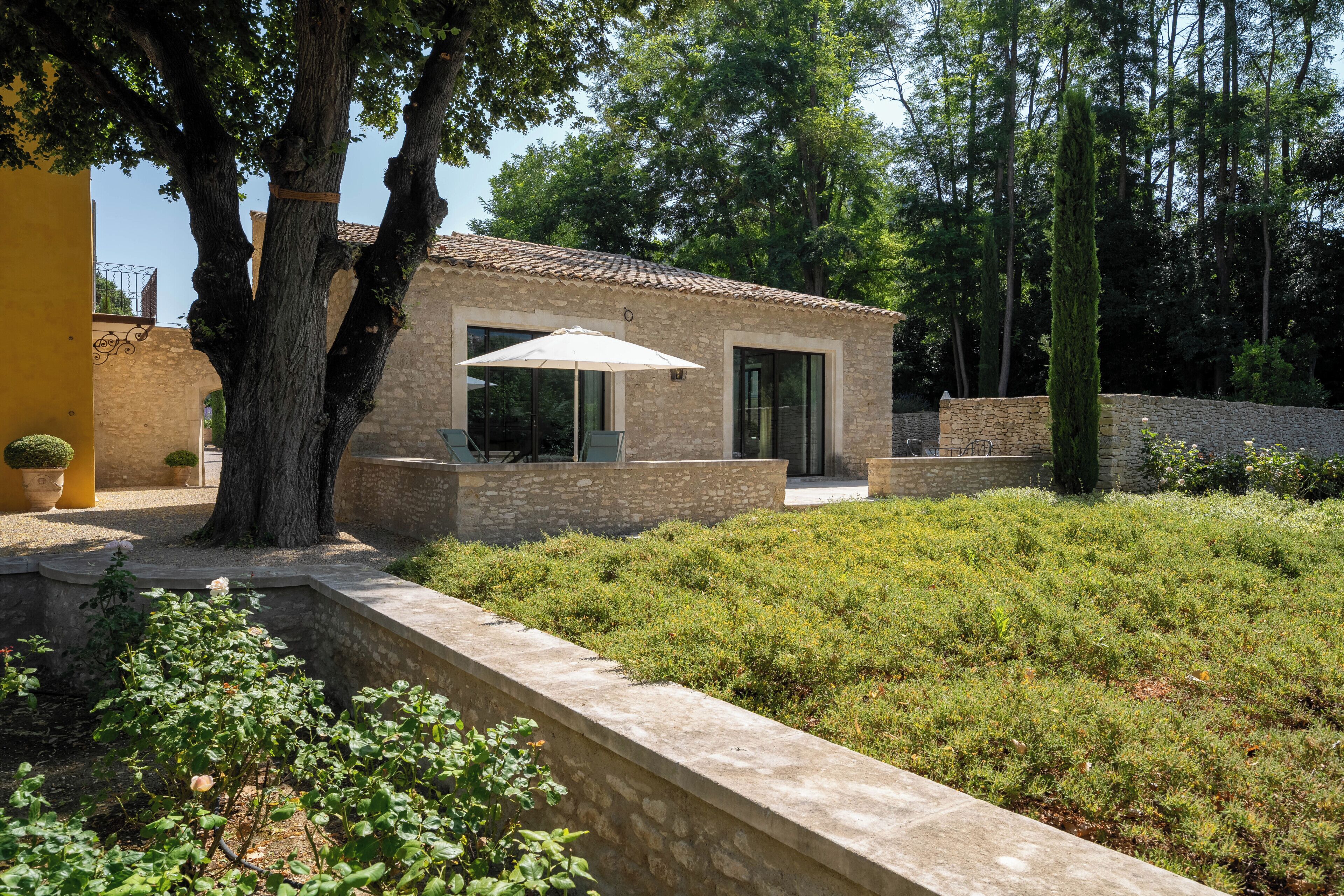 A stone building with a tiled roof, a patio with chairs and a table, and a garden with shrubs.