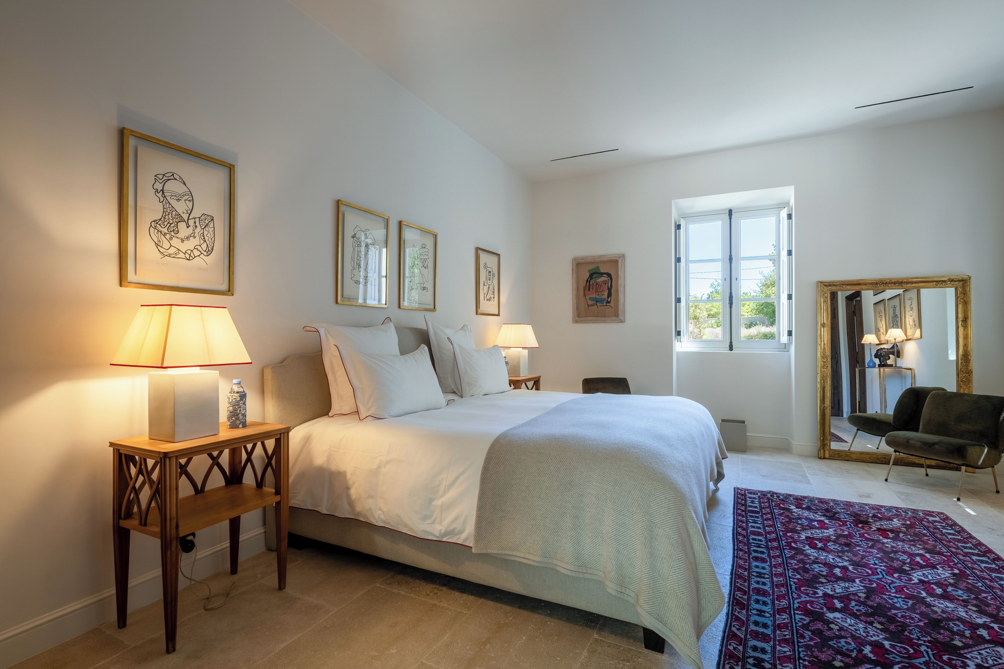 A bedroom with a bed, bedside lamps, a wooden nightstand, a chair, and framed artwork on the walls.