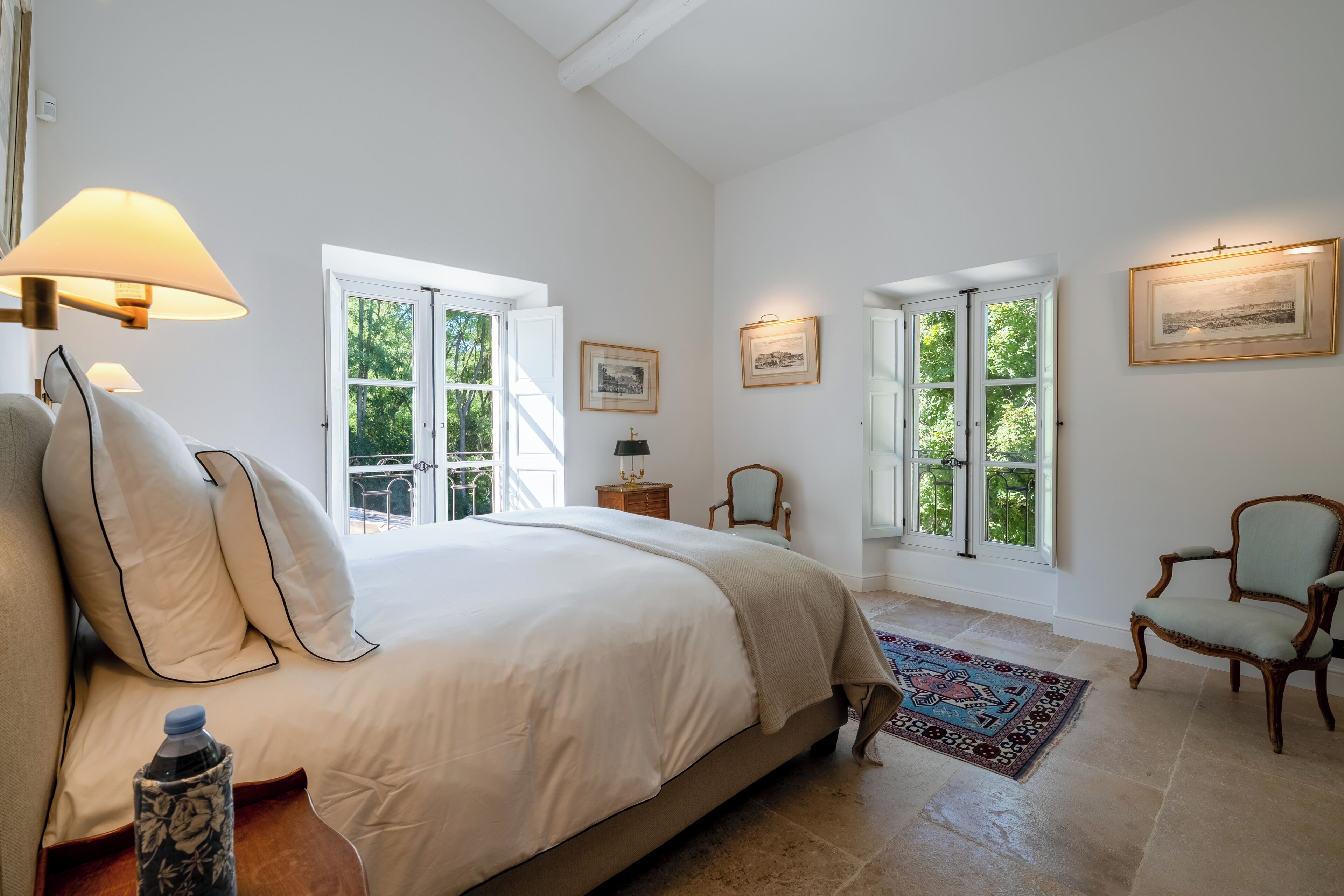 Een slaapkamer met een groot bed, twee ramen, een stoel en een vloerkleed.