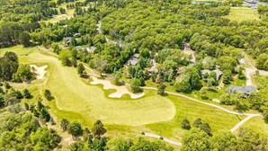 An aerial view of a golf course with a large putting green, surrounded by dense trees and residential properties.