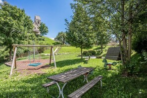 Une aire de jeux avec des balançoires et un toboggan, entourée d’arbres et agrémentée d’une table de pique-nique.