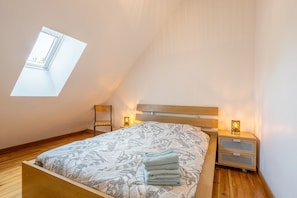Une chambre avec un plafond en pente, un puits de lumière, un lit recouvert d’une couvre-lit à motifs, deux tables de chevet avec des lampes et une tête de lit en bois.