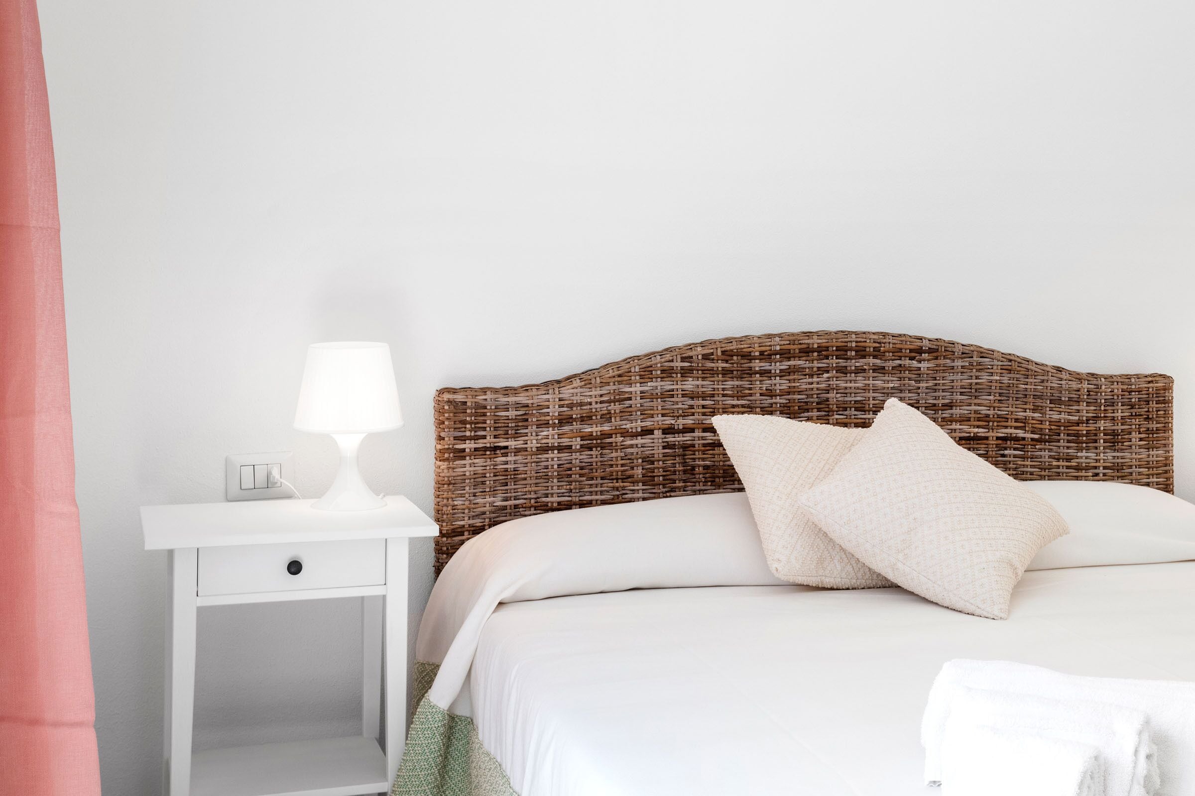 A neatly made bed with white bedding and two pillows, a white bedside table with a lamp, and a wicker headboard.