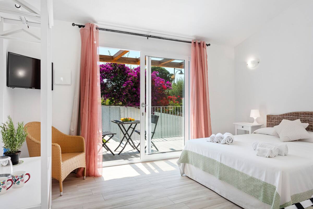A bedroom with a bed, a TV, a balcony with a table and chairs, and a view of flowers.