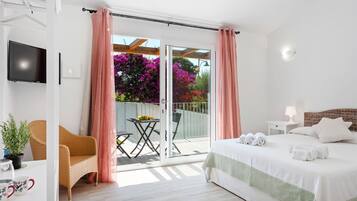 A bedroom with a bed, a TV, a balcony with a table and chairs, and a view of flowers.