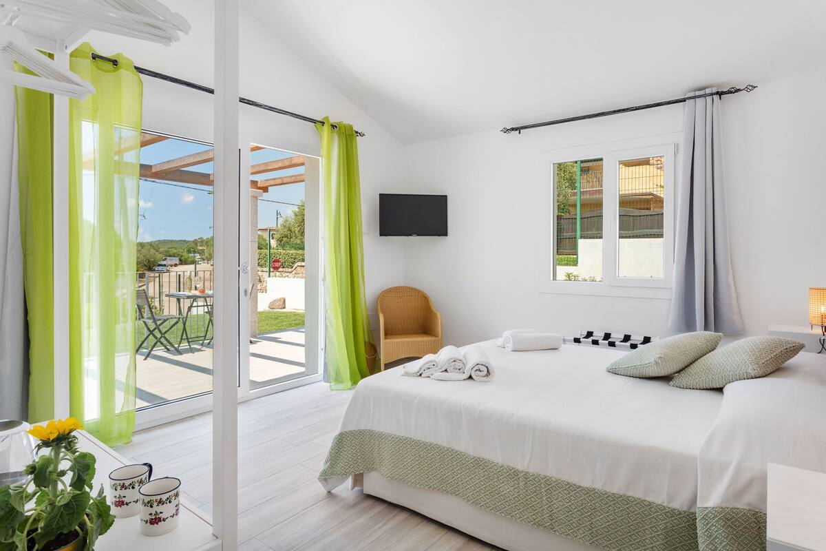 A modern bedroom with a large bed, a flat-screen TV, a wicker chair, and a view of the outdoors.