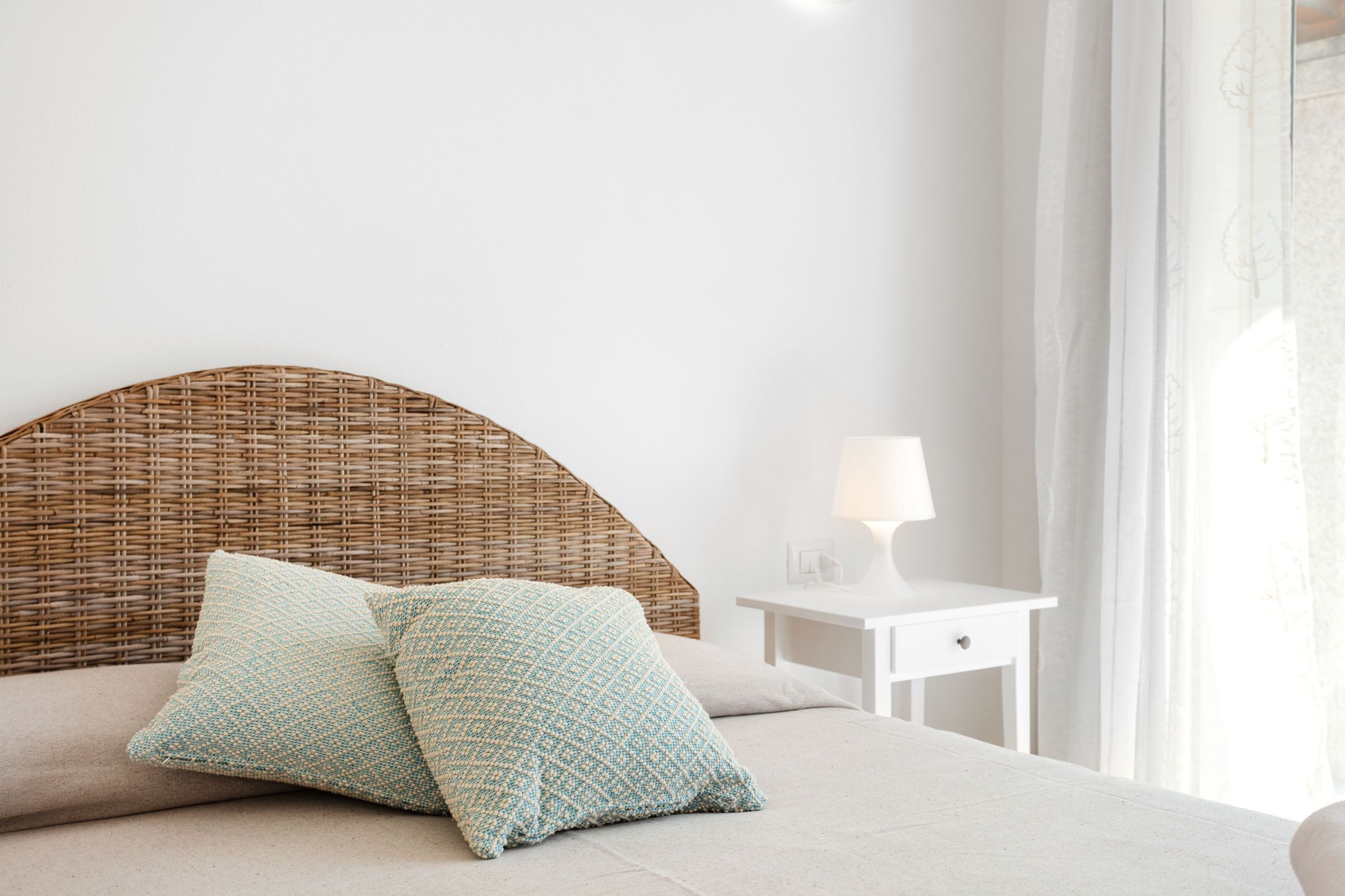 A bedroom with a wicker headboard, two pillows, a bedside table with a lamp, and a window with curtains.