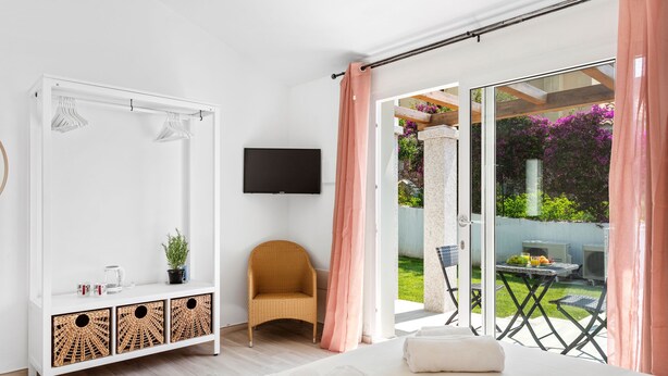 A modern bedroom with a large bed, a white wardrobe, a flat-screen TV, a wicker chair, and a balcony with a view of a garden and outdoor furniture. - Sardegna è - New rooms (Olbia)