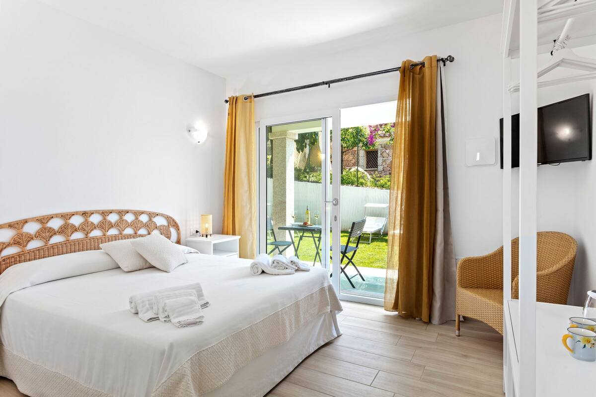 A hotel room with a bed, a glass door leading to an outdoor area, a wicker chair, and a TV mounted on the wall.