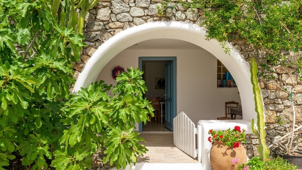 Une entrée en arc surmontée d’un mur de pierre, avec une porte blanche, donnant sur une pièce où l’on aperçoit une table et des chaises.