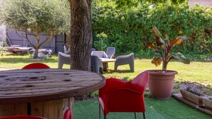 A wooden table with chairs, a potted plant, and a grassy area with a tree and bushes.