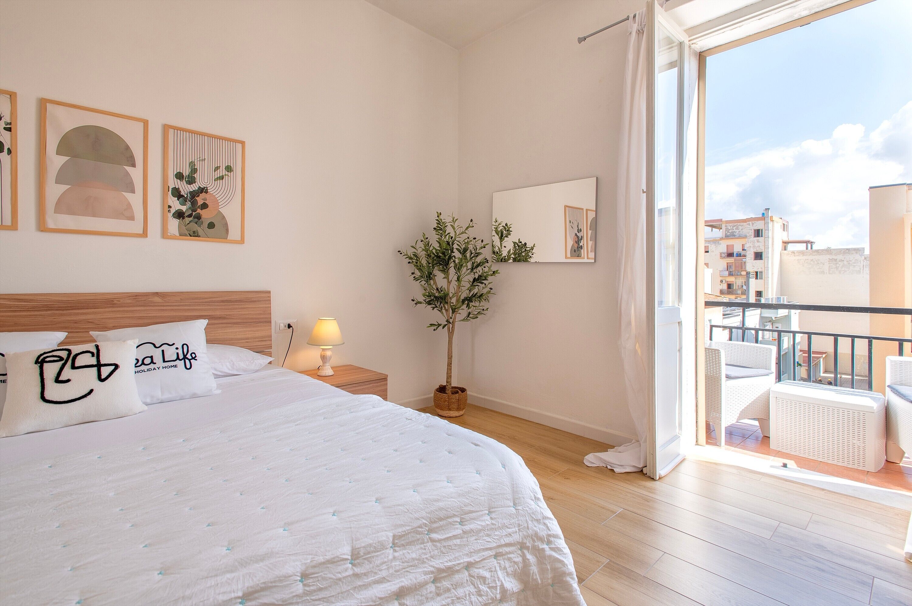 A bedroom with a bed, pillows, a nightstand, a lamp, a potted plant, a mirror, and a balcony with outdoor furniture.